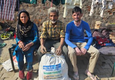 Three individuals sitting outside with a Jan Sewa Trust food kit