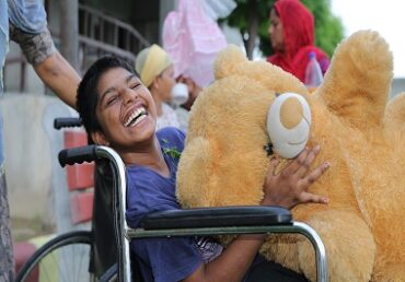 Child in wheelchair holding large teddy bear, smiling at camera